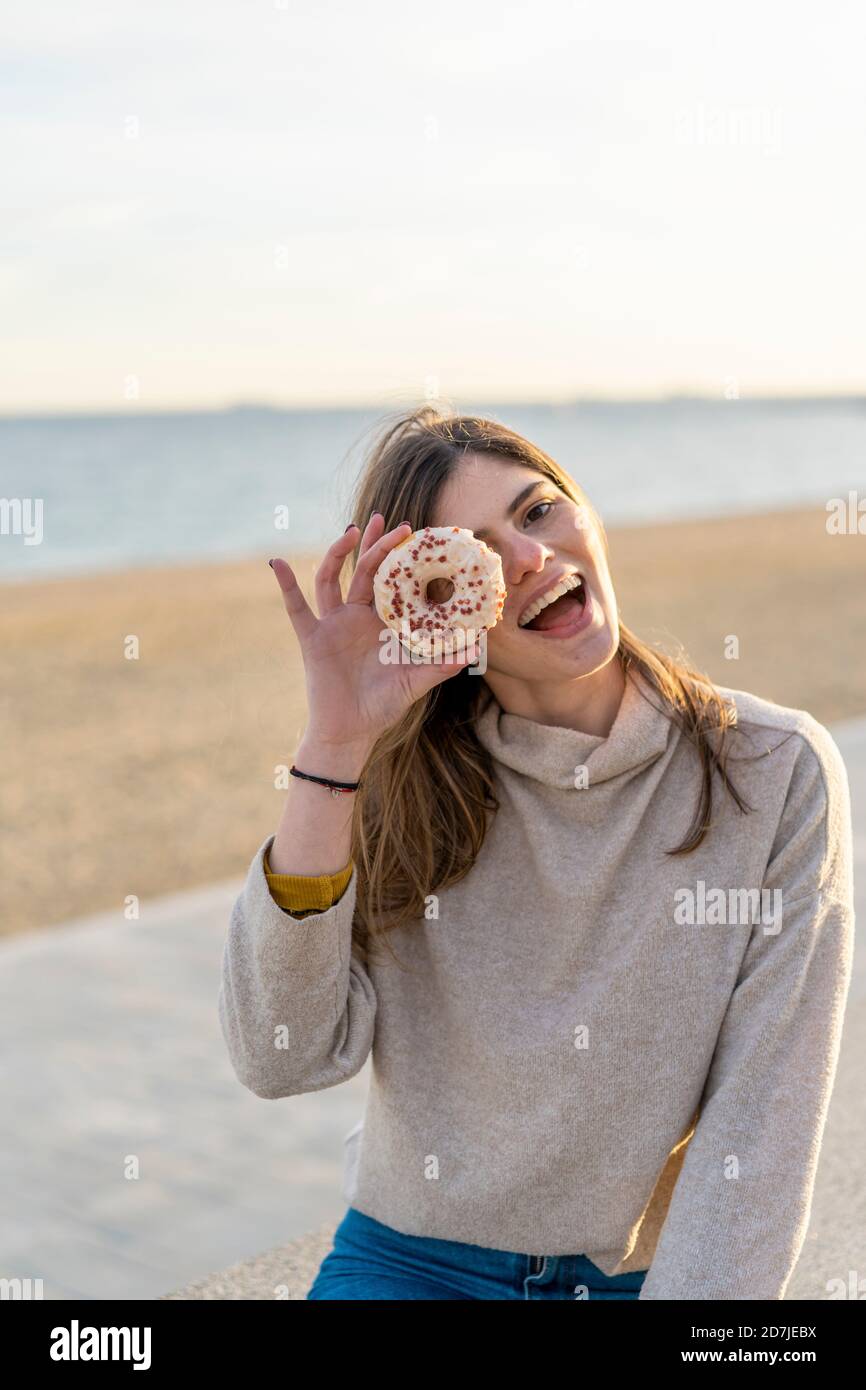 Cheerful young woman holding fresh donut in front of face while sitting at beach during sunset Stock Photo