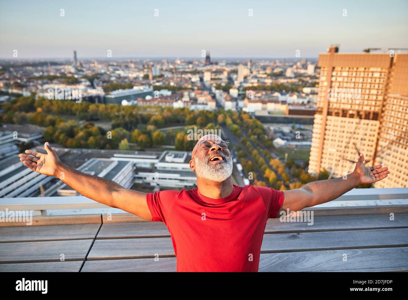 Happy bearded man with arms outstretched leaning back while standing on building terrace in city during sunset Stock Photo