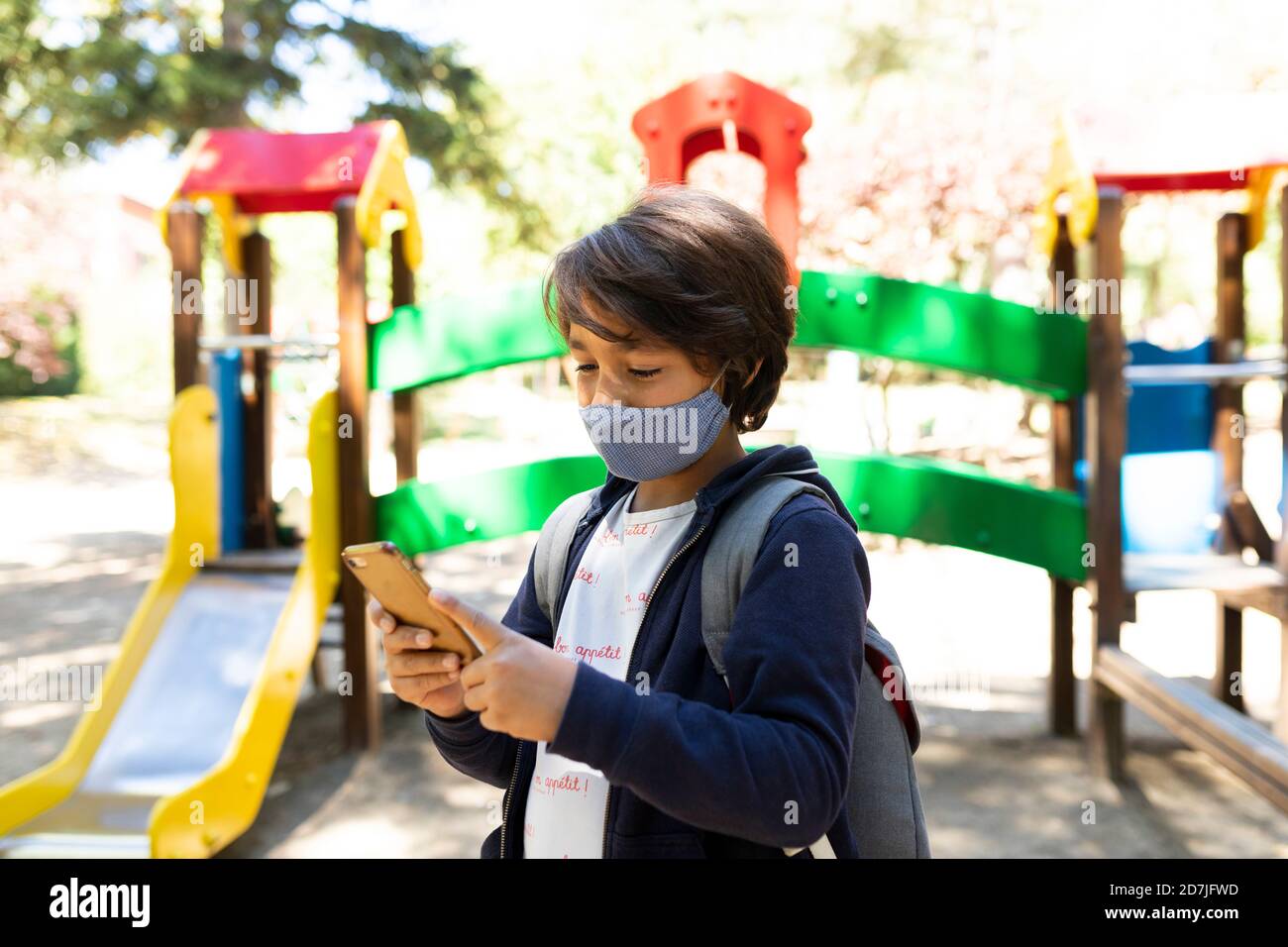Schoolboy wearing mask using smart phone while standing in schoolyard Stock Photo