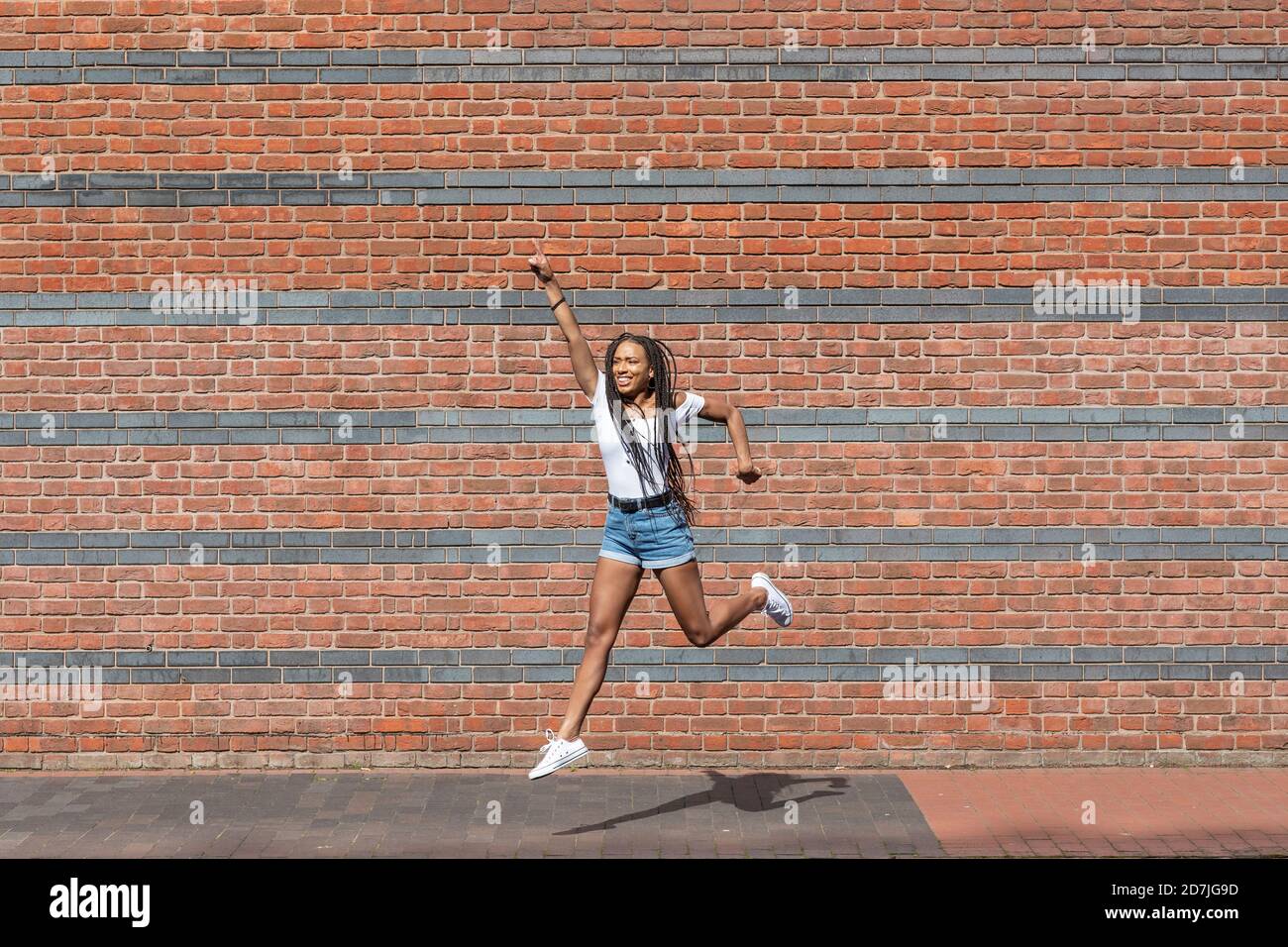 Young woman smiling while jumping against brick wall Stock Photo