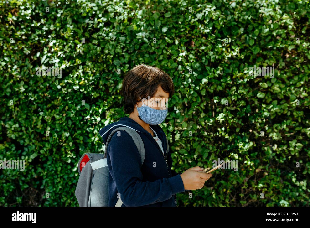 Schoolboy wearing mask using smart phone while standing against plants Stock Photo