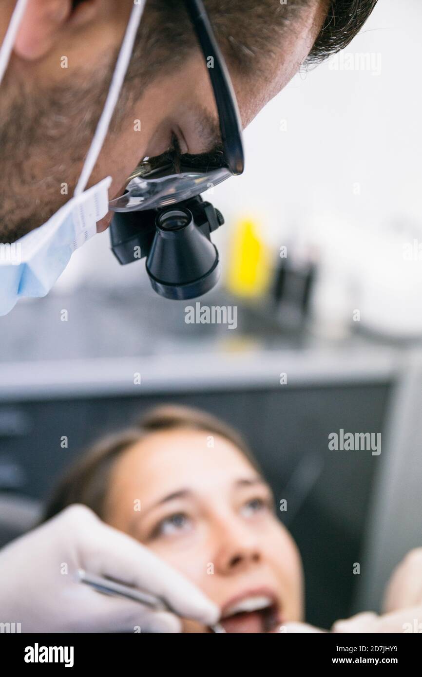 Male dentist in surgical loupes examining teeth of female patient in
