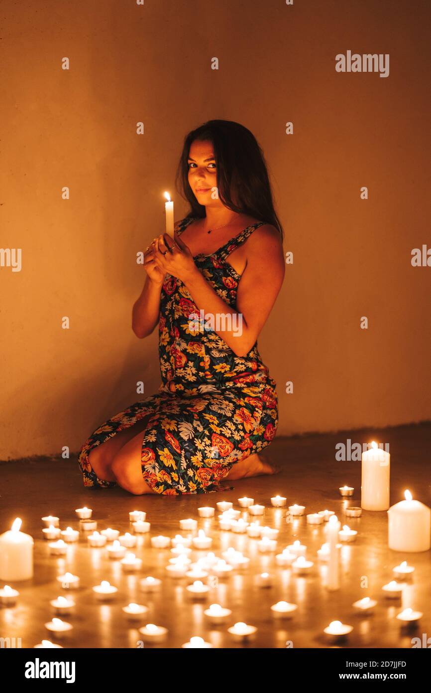 Woman with illuminated candles kneeling on floor by wall in darkroom Stock Photo