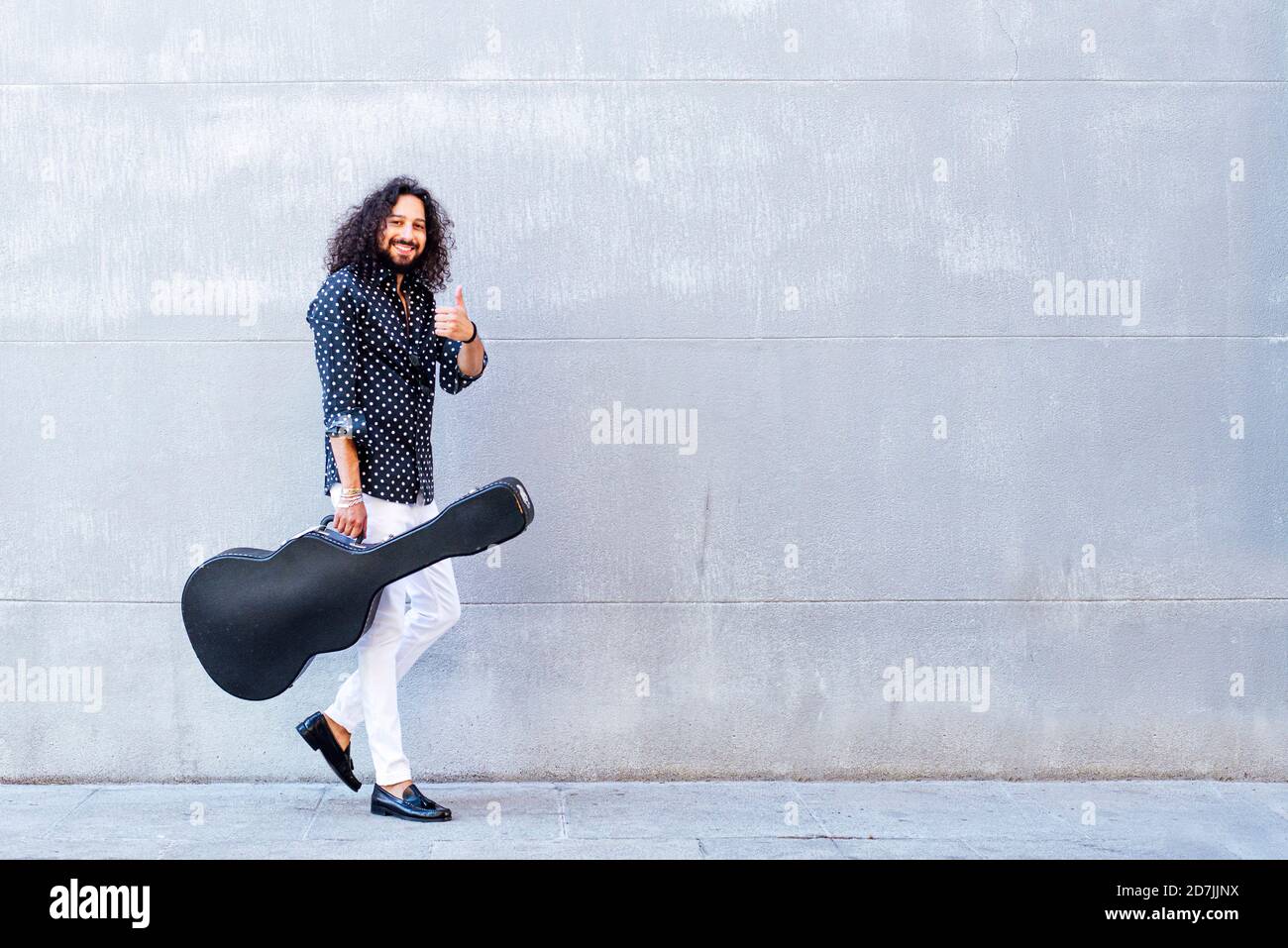 Smiling man with guitar showing thumps up while walking against gray wall Stock Photo