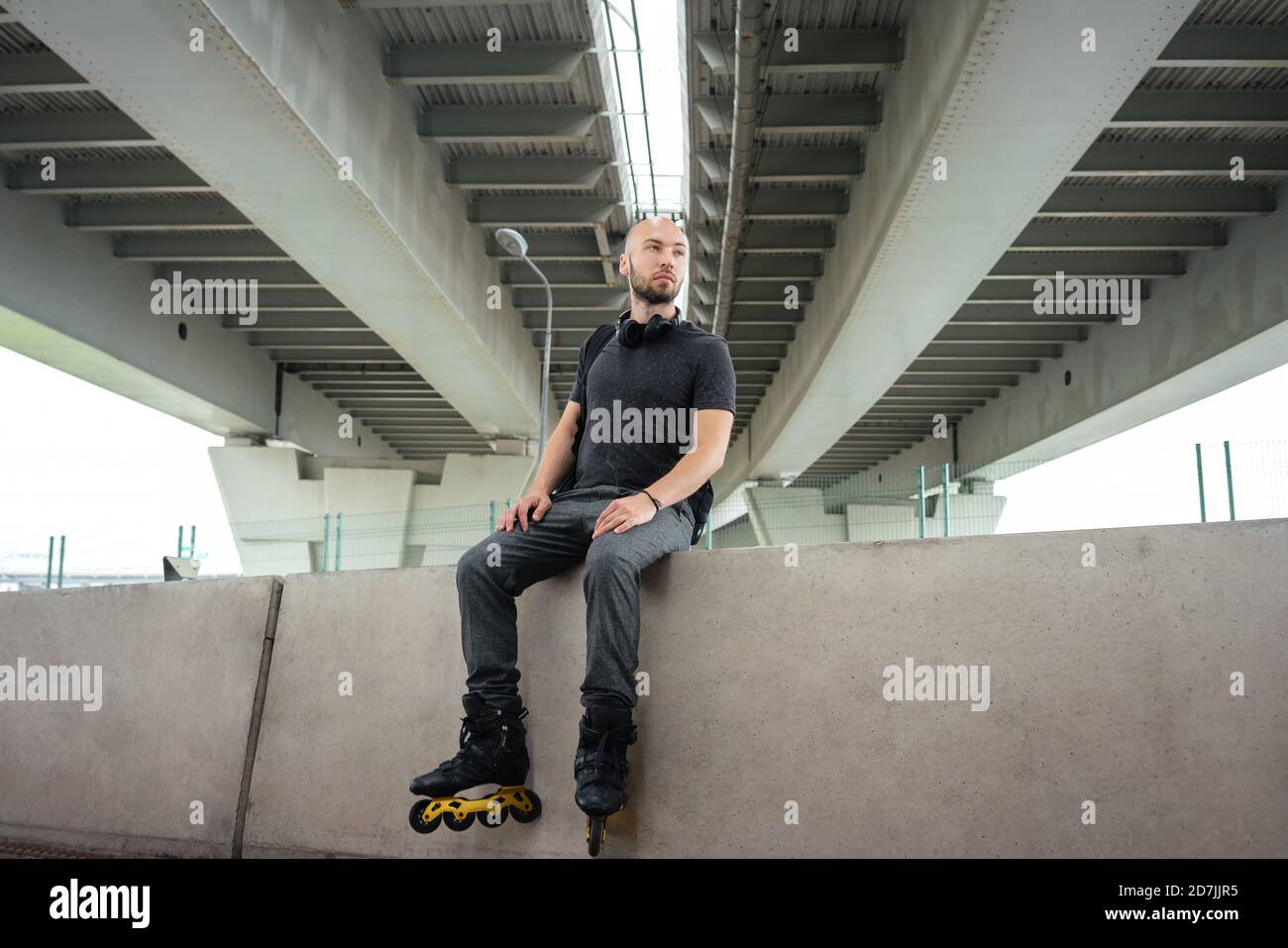 Thoughtful young man wearing inline skates while sitting on retaining wall under bridge Stock Photo