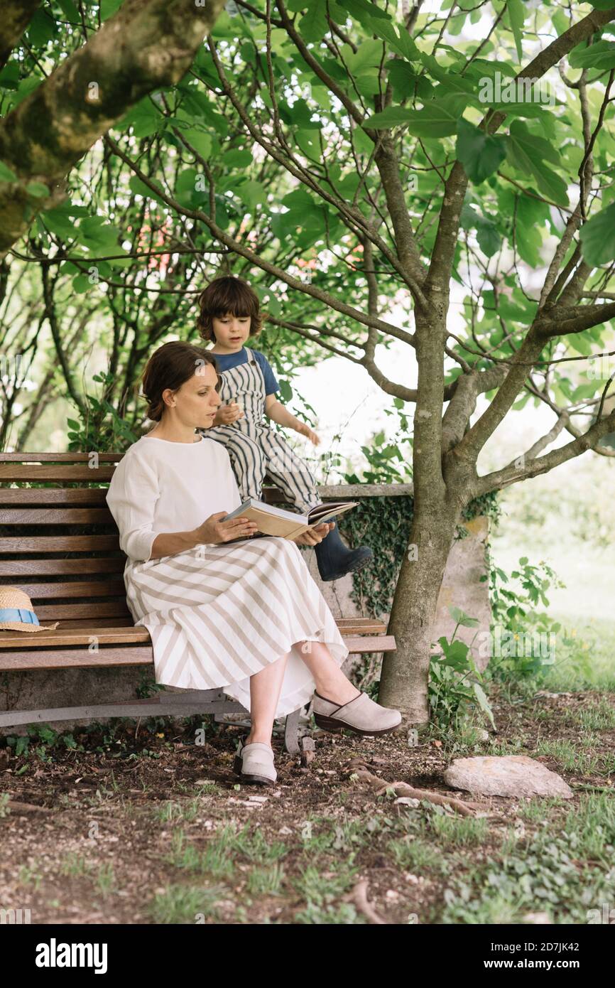 Beautiful woman and boy reading book while sitting on bench at backyard ...