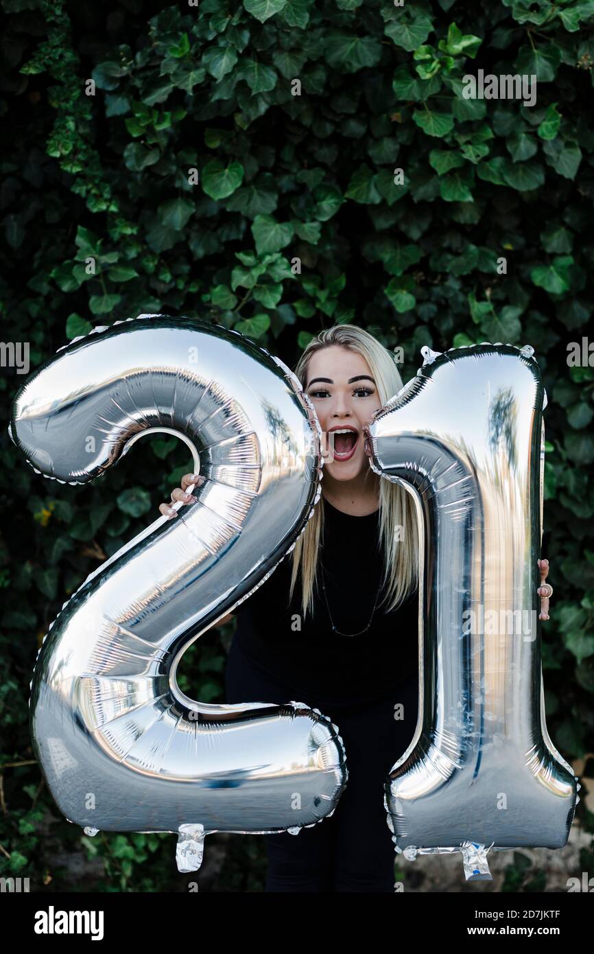 Young woman with mouth open holding number 21 balloons while standing against plants in park Stock Photo
