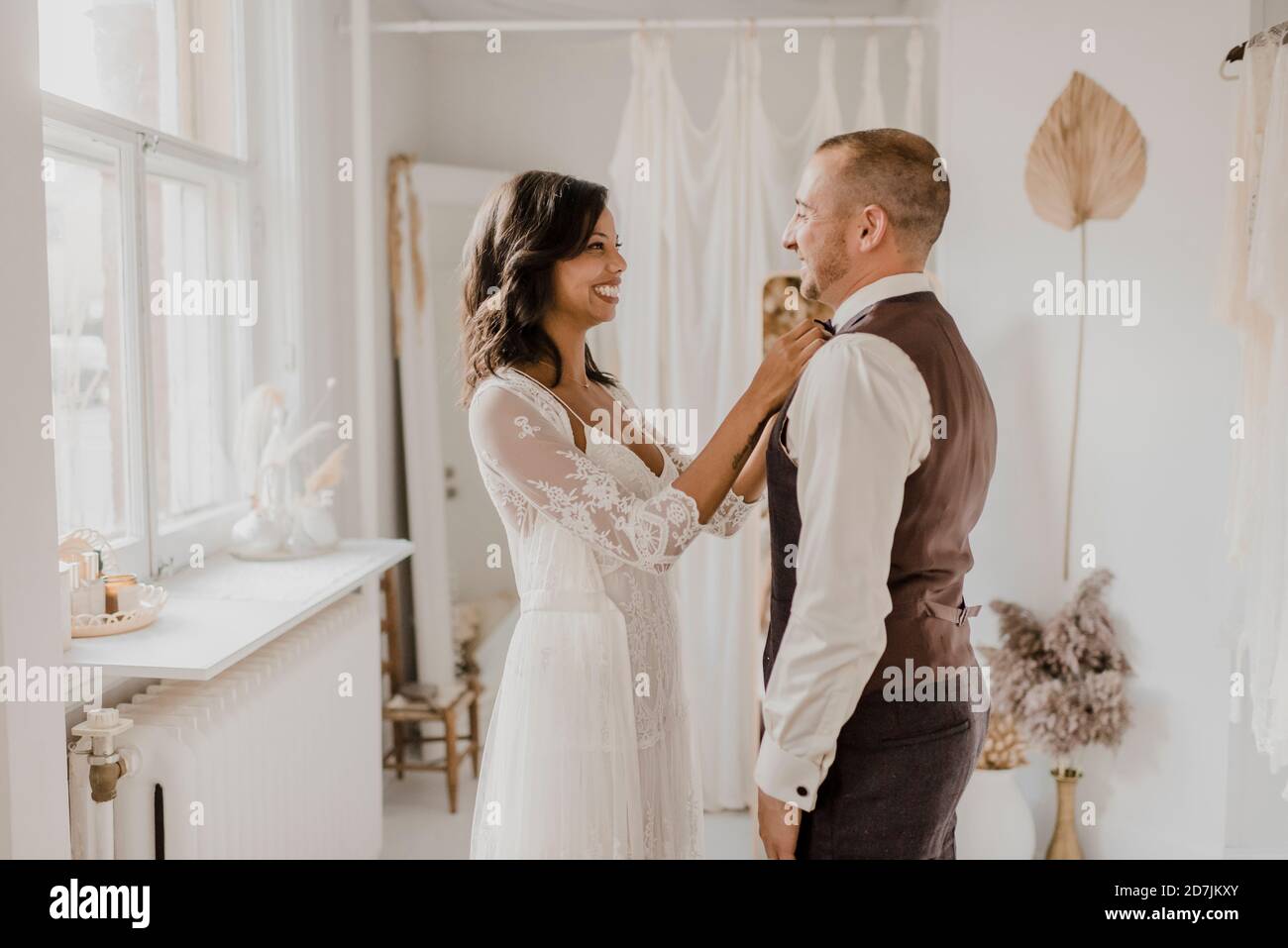 Smiling bride tying bow to bridegroom at wedding dress shop Stock Photo