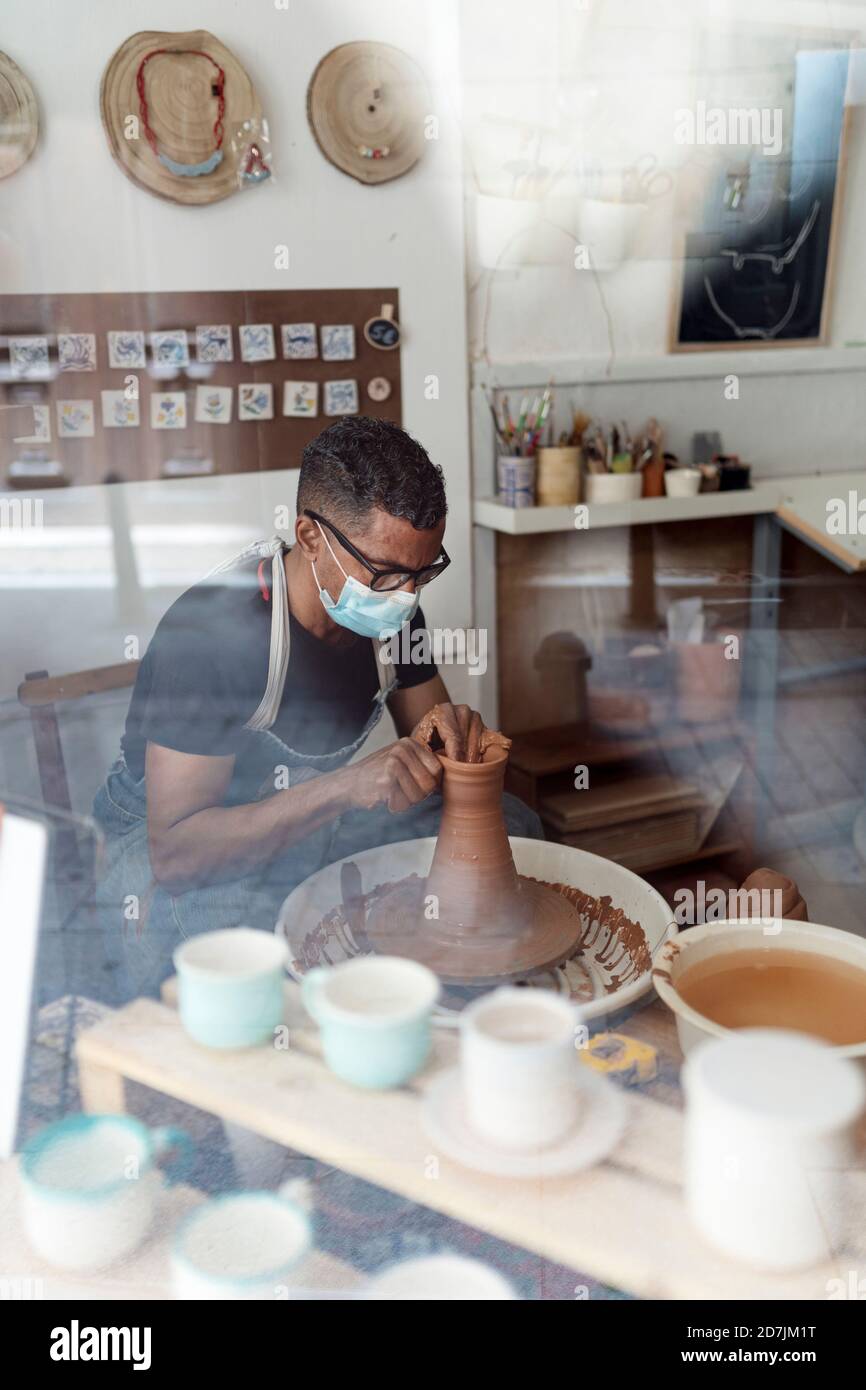 Male potter wearing mask molding shape to clay in workshop seen through window Stock Photo