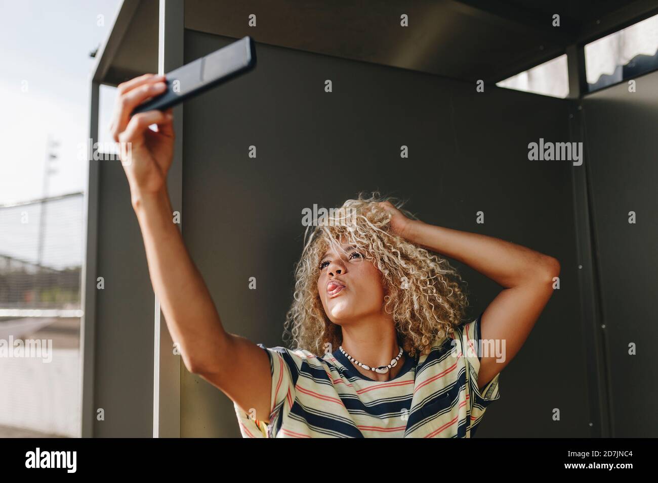 Woman Sticking Out Tongue While Taking Selfie Through Smart Phone Against Metallic Wall Stock 