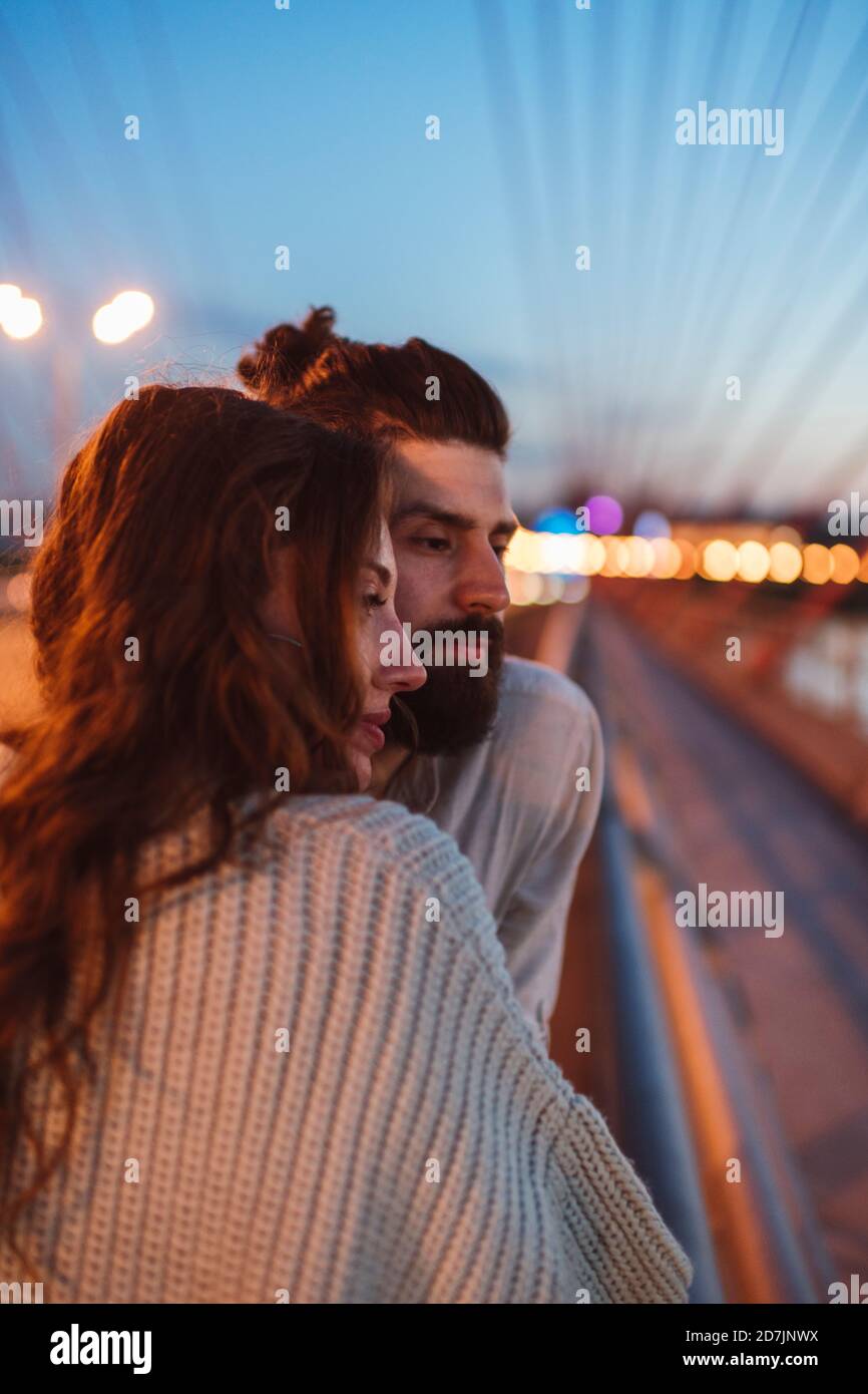 Thoughtful couple looking away while standing on bridge in city at dusk Stock Photo