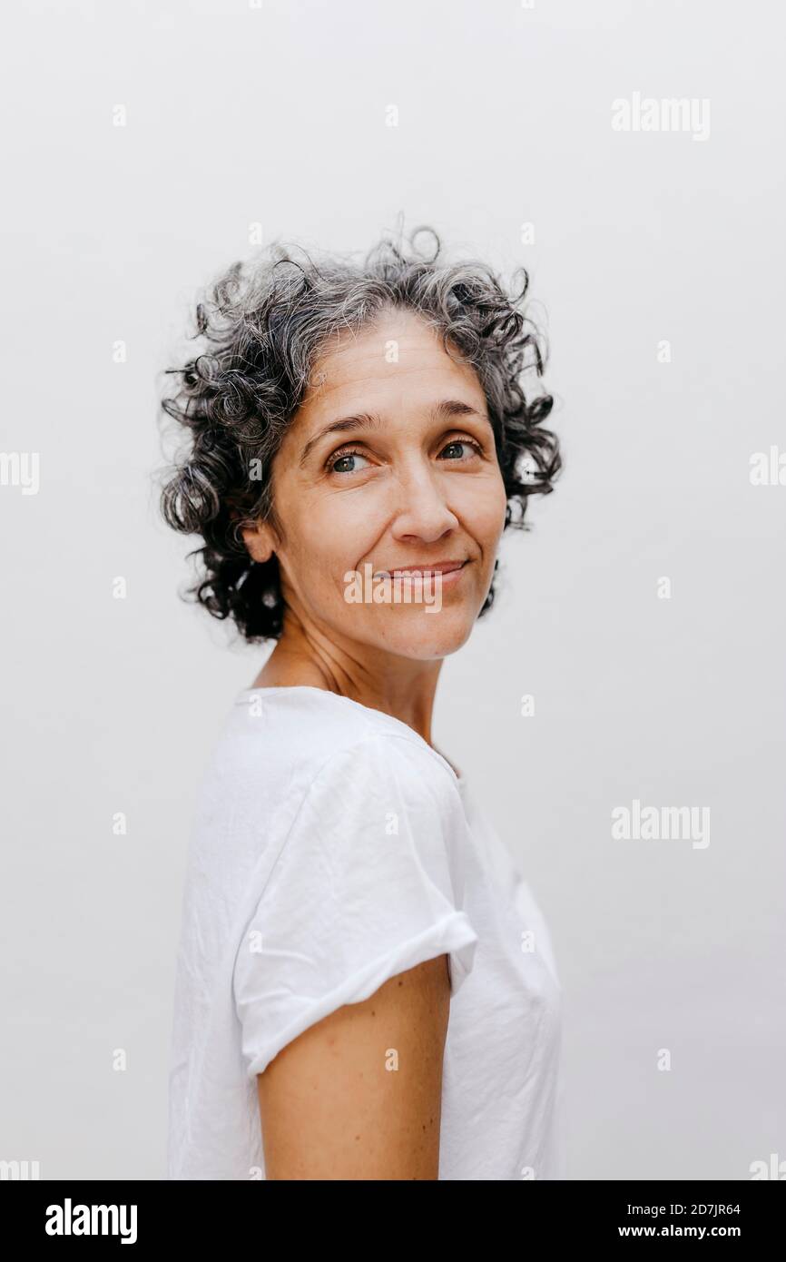 Mature woman with short curly hair looking away while standing against white wall Stock Photo