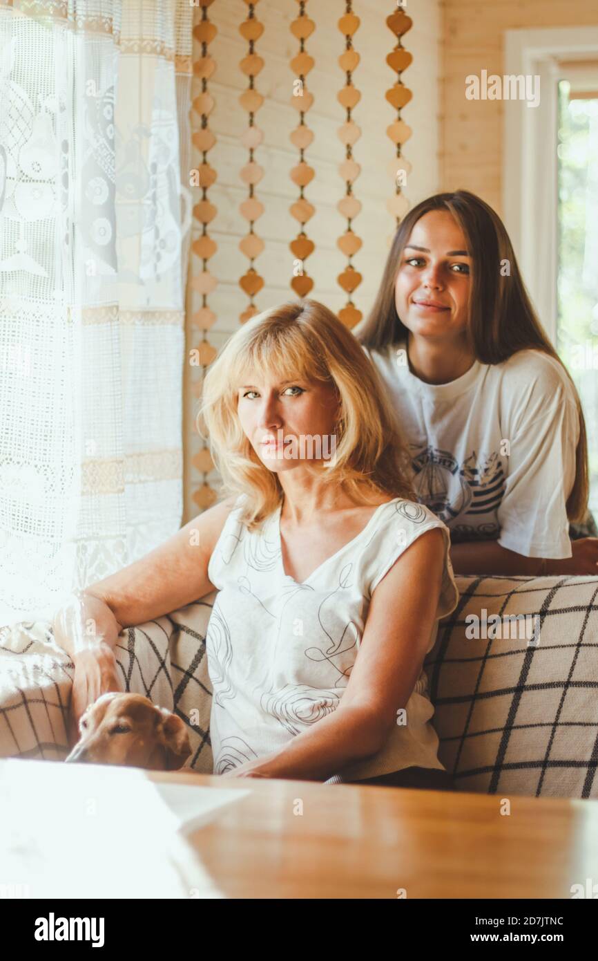 Loving adult 20s daughter hug elderly mother from behind while mom sitting on couch people posing looking at camera smiling feels happy, concept of mu Stock Photo