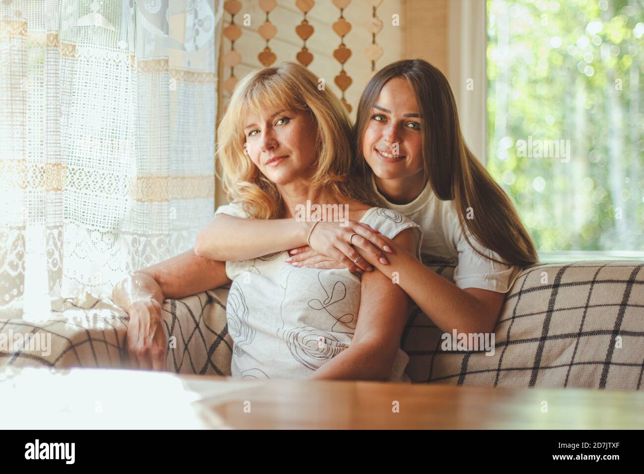 Loving adult 20s daughter hug elderly mother from behind while mom sitting on couch people posing looking at camera smiling feels happy, concept of mu Stock Photo
