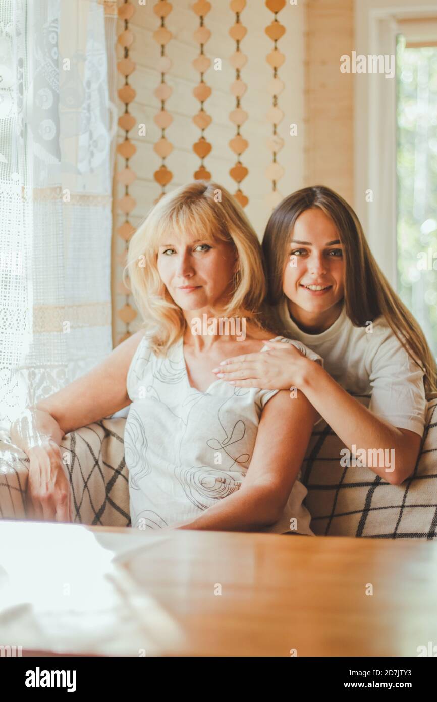 Loving adult 20s daughter hug elderly mother from behind while mom sitting on couch people posing looking at camera smiling feels happy, concept of mu Stock Photo