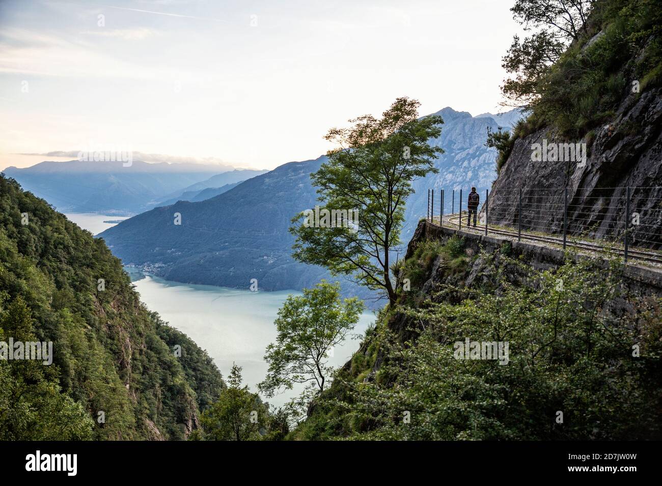 Italy, Province of Sondrio, Silhouette of hiker admiring Lake Mezzola from fenced edge of Tracciolino trail Stock Photo