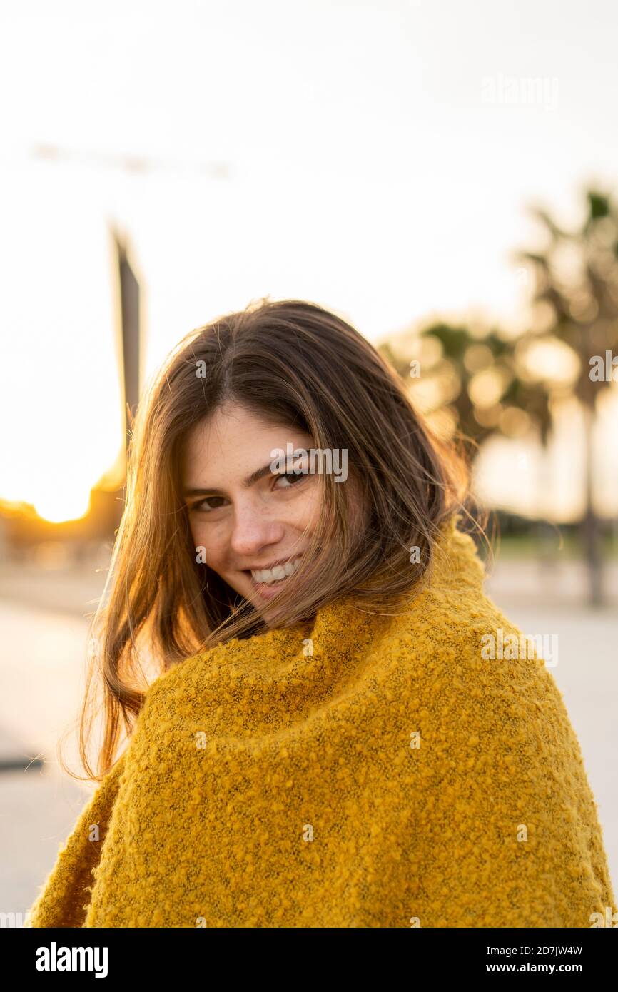 Smiling beautiful young woman wrapped in brown jacket at beach during sunset Stock Photo