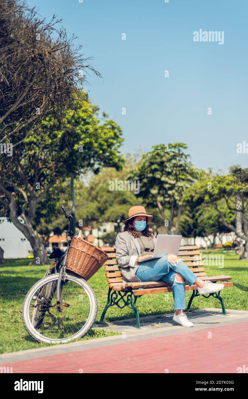 Vertical image of a female blogger typing on her laptop outdoors while wearing a medical mask and riding a bicycle. New normal concept. Copy space Stock Photo