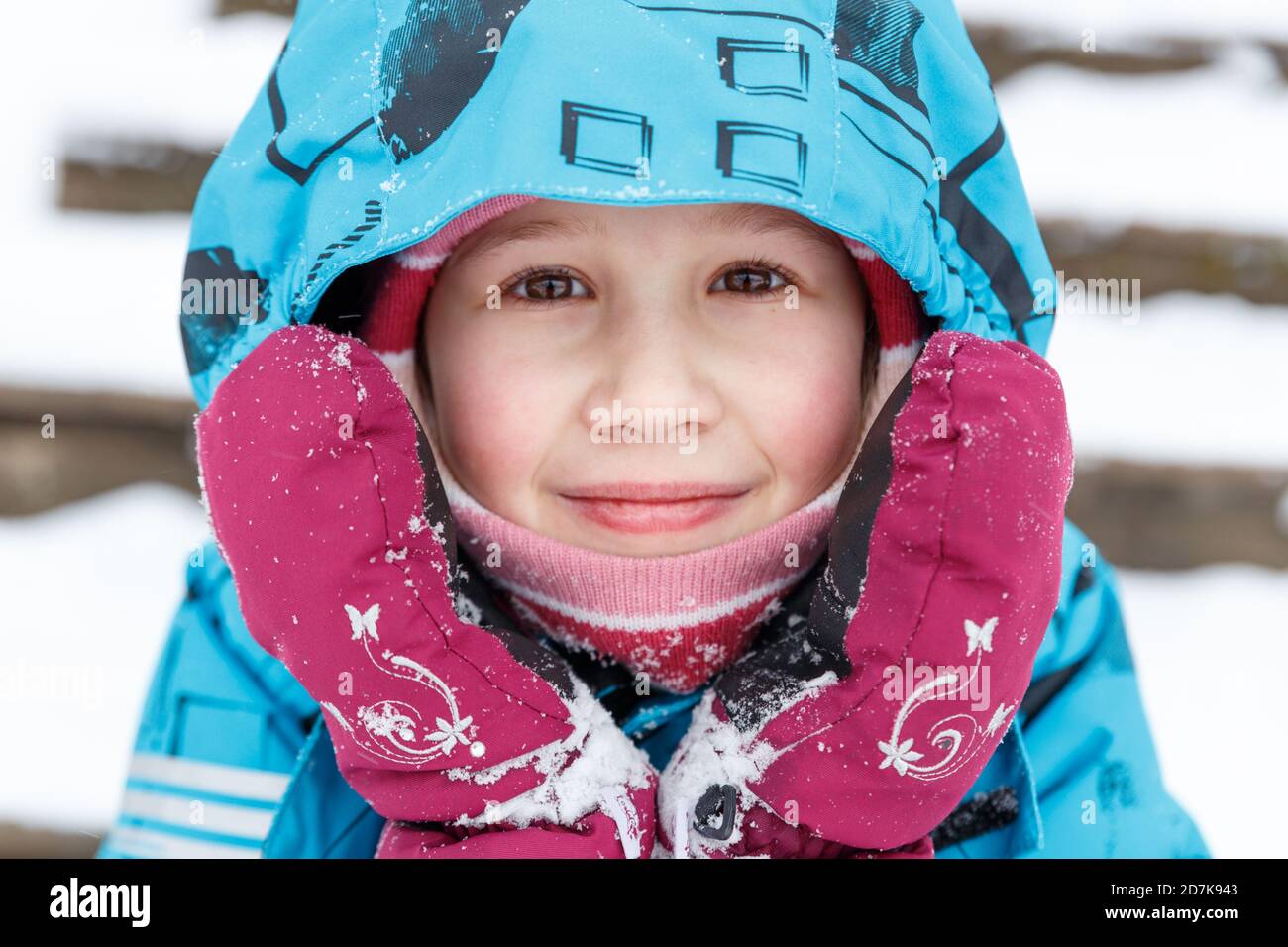 Close-up portrait of cute caucasian baby in winter clothes. A 6-7 year old girl with ruddy cheeks from frost, in winter overalls and mittens, looks at Stock Photo