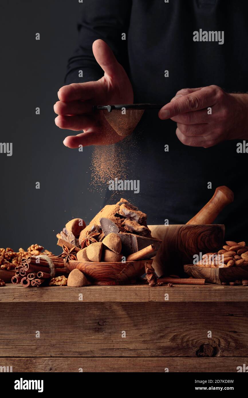 The cook sprinkles the chocolates with cocoa powder. On an old wooden table candy, coffee beans, cinnamon, anise, nuts, and pieces of broken black choco Stock Photo