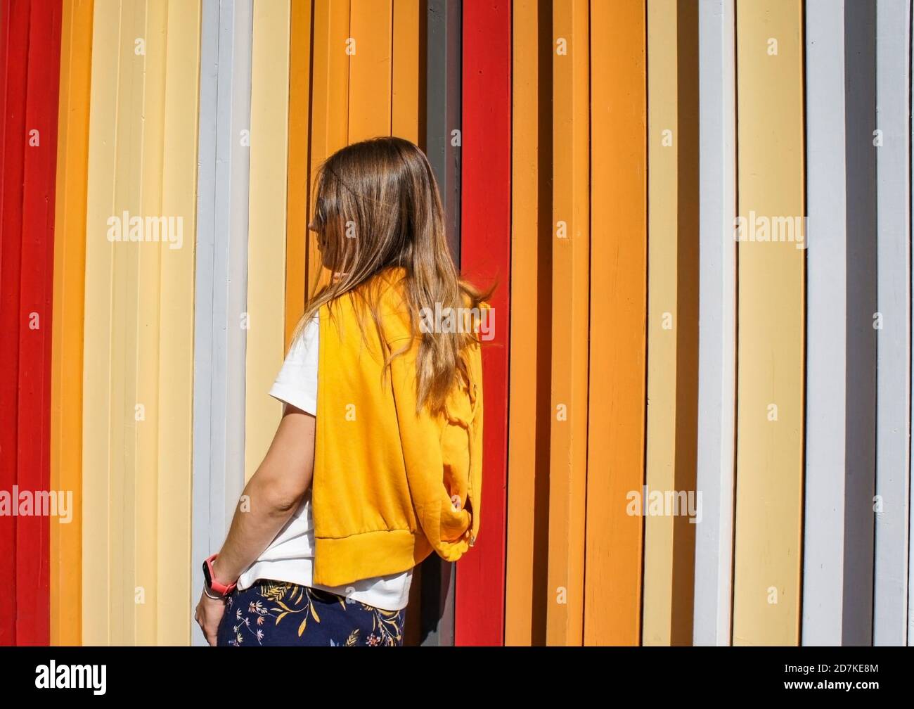 Young blond woman from the back in a yellow sweatshirt stands against the background of a bright colored striped wall. Caucasian teenager girl Stock Photo