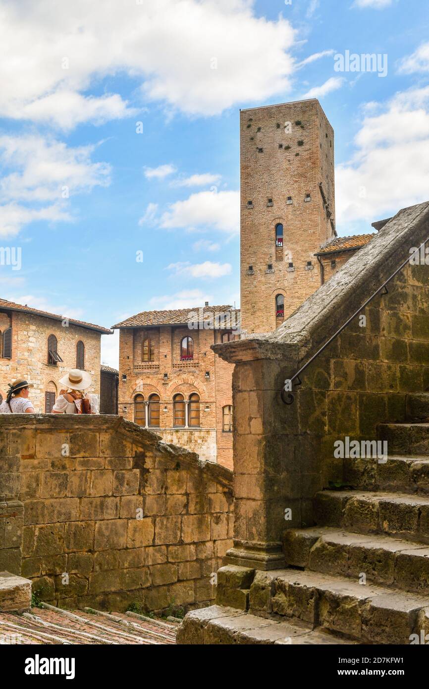 Tourist women with hats on the stairs of the Duomo of San Gimignano admiring the main square with the medieval Torre Chigi, Siena, Tuscany, Italy Stock Photo