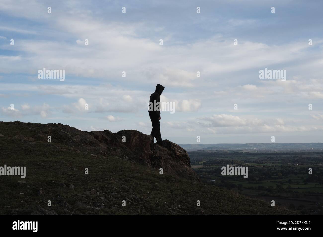 A hooded figure walking towards the top of a rocky outcrop. Looking down across the landscape Stock Photo