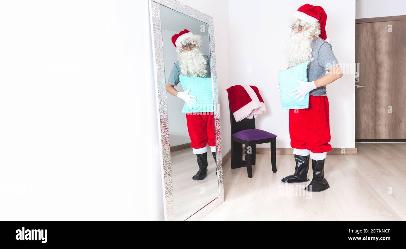Man dressing up as Santa Claus, putting a pillow on his belly to look fatter, looking at himself in the mirror in the living room Stock Photo