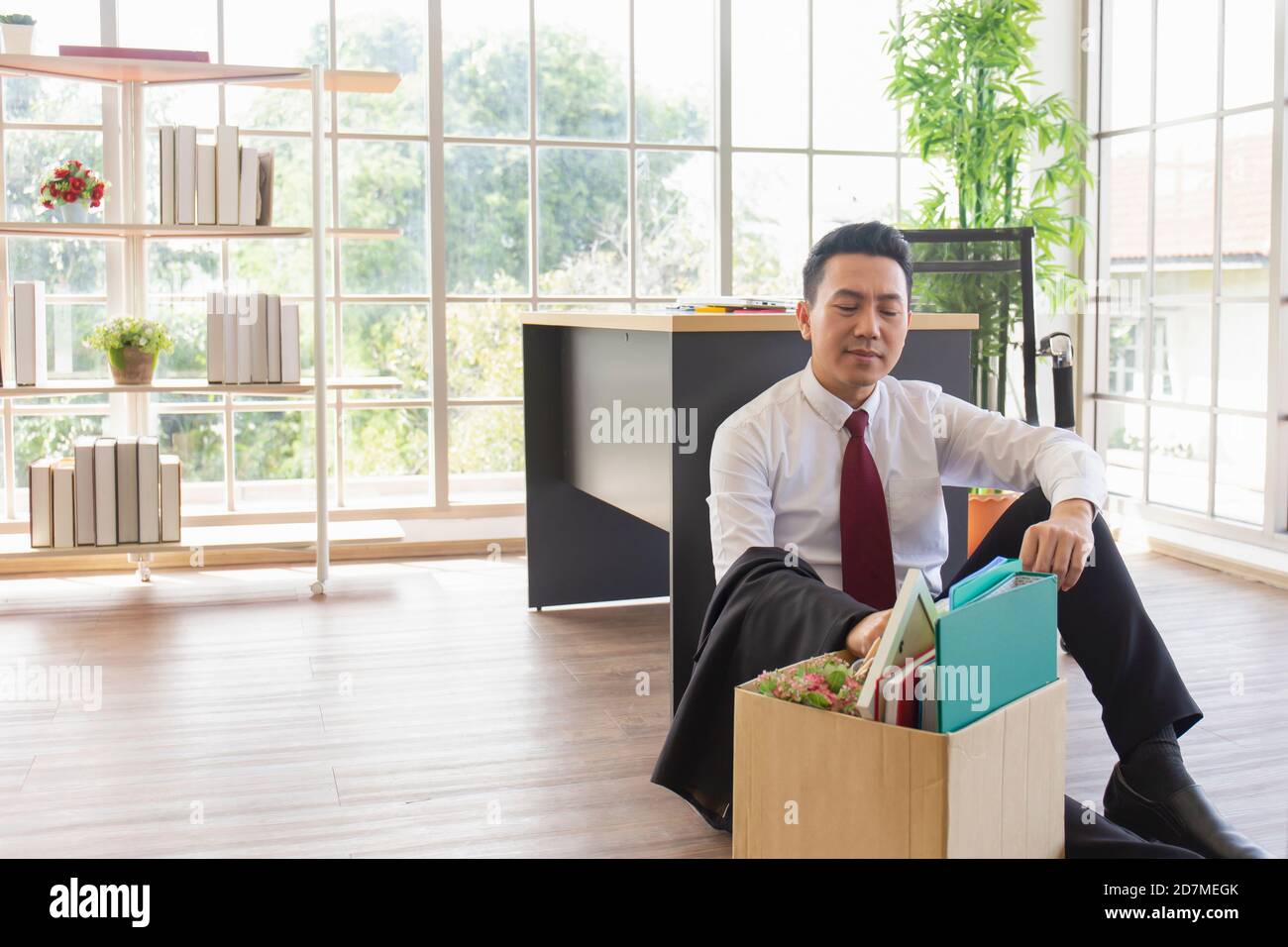 An Asian male company worker sits on the floor beside a desk with a cardboard box containing his personal belongings. Stock Photo