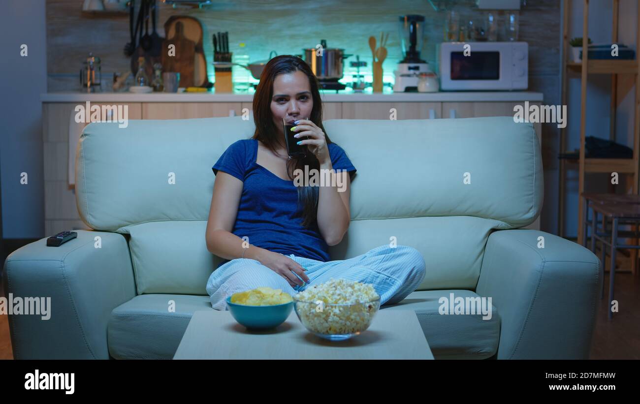 Happy woman spending her free time watching TV eating popcorn and drinking juice. Excited amused home alone lady enjoying the evening at home sitting on comfortable couch dressed in pajamas. Stock Photo