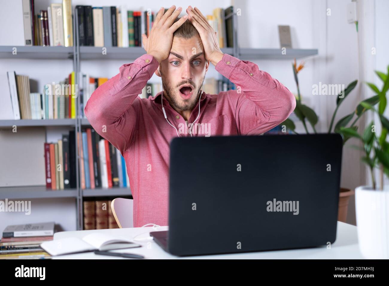 Attractive young man sitting at the table at home working with a laptop on a calling and feeling surprise Stock Photo