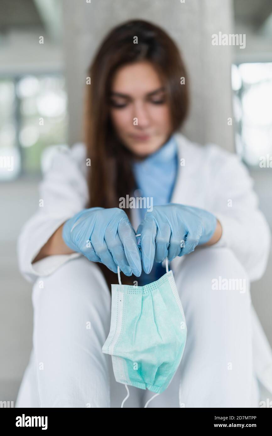 Sad and hopeless young female doctor holding protective mask sitting on floor at hospital after shift. Coronavirus covid-19 concept. Stock Photo