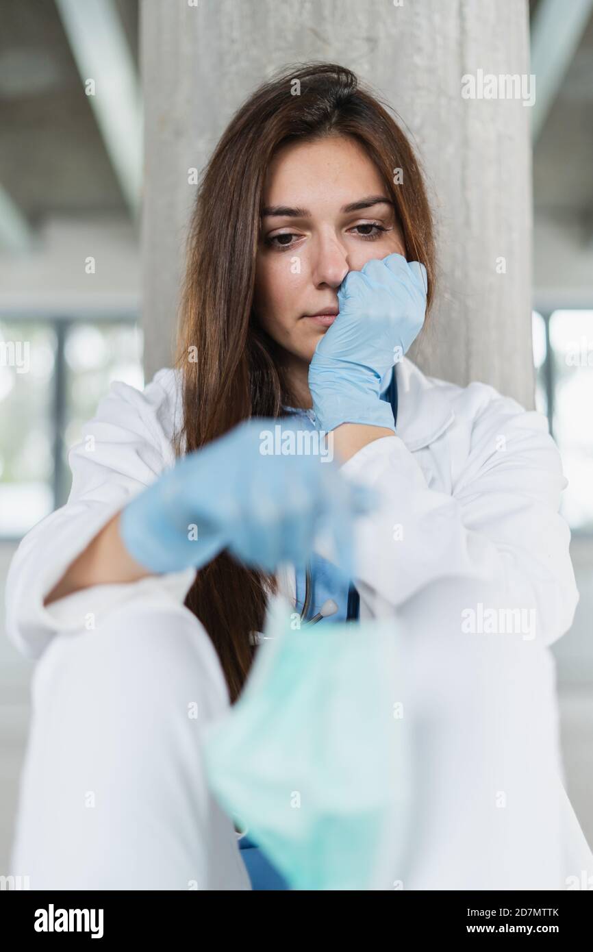 Sad and hopeless young female doctor holding protective mask sitting on floor at hospital after shift. Coronavirus covid-19 concept. Stock Photo
