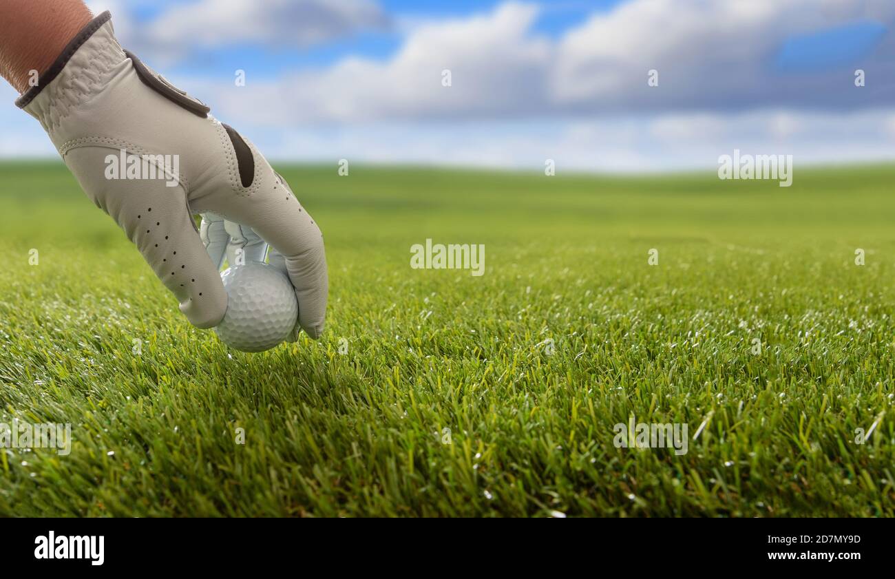 Golfer hand in a white glove holding a golfball, green course lawn background, close up view. Blue cloudy sky background. Stock Photo