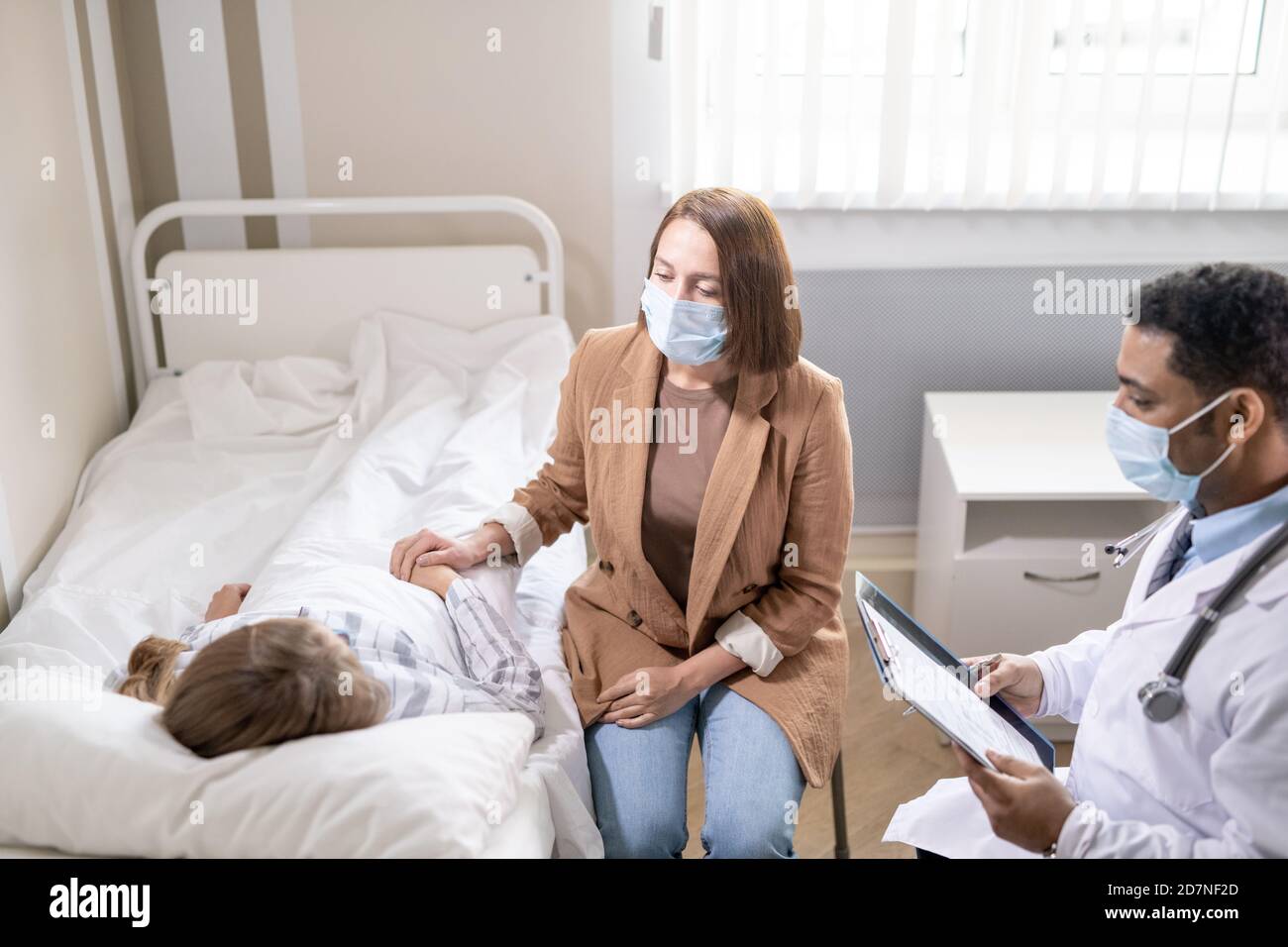 Young woman in protective mask sitting by bed of sick friend in covid hospital Stock Photo
