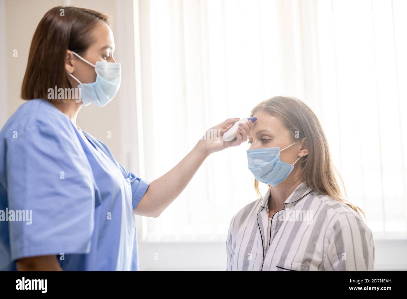 Young nurse in blue uniform and mask holding thermometer by patient forehead Stock Photo