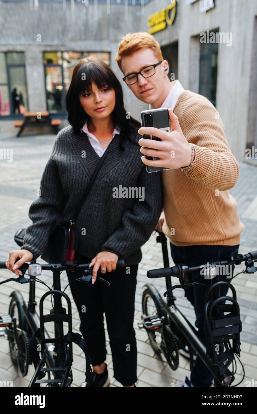 Unusual couple taking selfie on the street, tugging along ebikes Stock Photo
