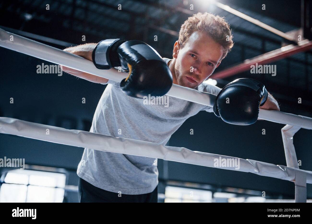 Tired young man in white shirt and protective gloves leaning on knots of boxing ring Stock Photo