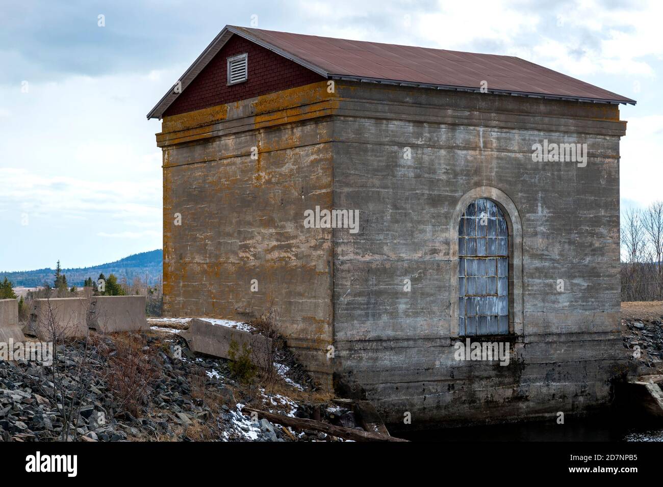 An antique hydro-electric generating station. Moss grows on faded concrete sides. Arched window facing water. Overcast sky. Station is no longer in us Stock Photo