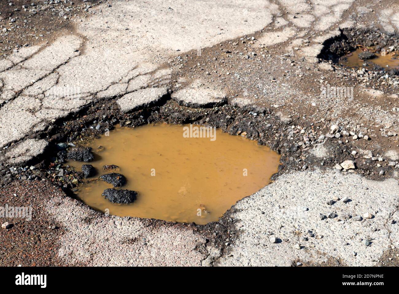 Potholes on a paved road. The road is in very poor condition. There is dirty water in the potholes. Stock Photo