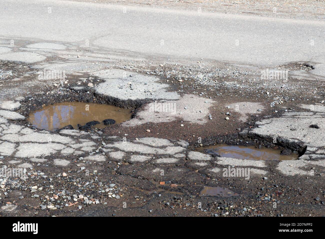 Potholes on a paved road. The road is in very poor condition. There is dirty water in the potholes. Stock Photo