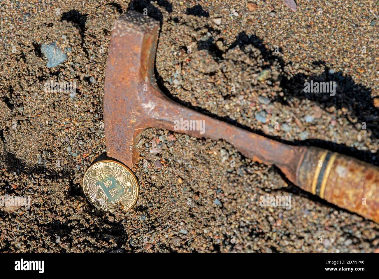 A pick axe digging a bitcoin out of sandy ground. The point of the pick axe is buried behind a single golden bitcoin. Stock Photo