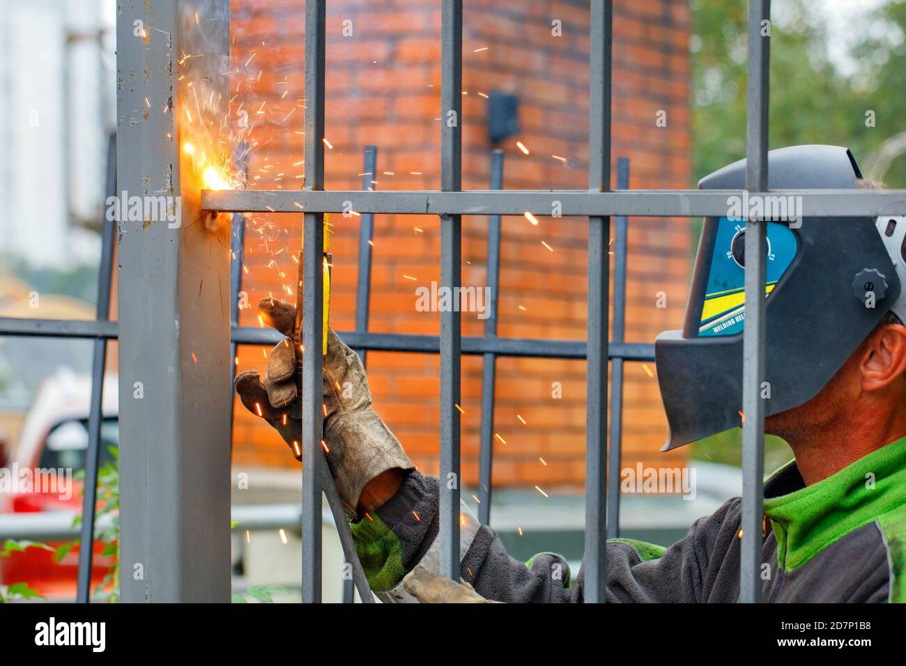 A welder wearing a protective helmet and gloves is welding a metal fence around an apartment building. Stock Photo