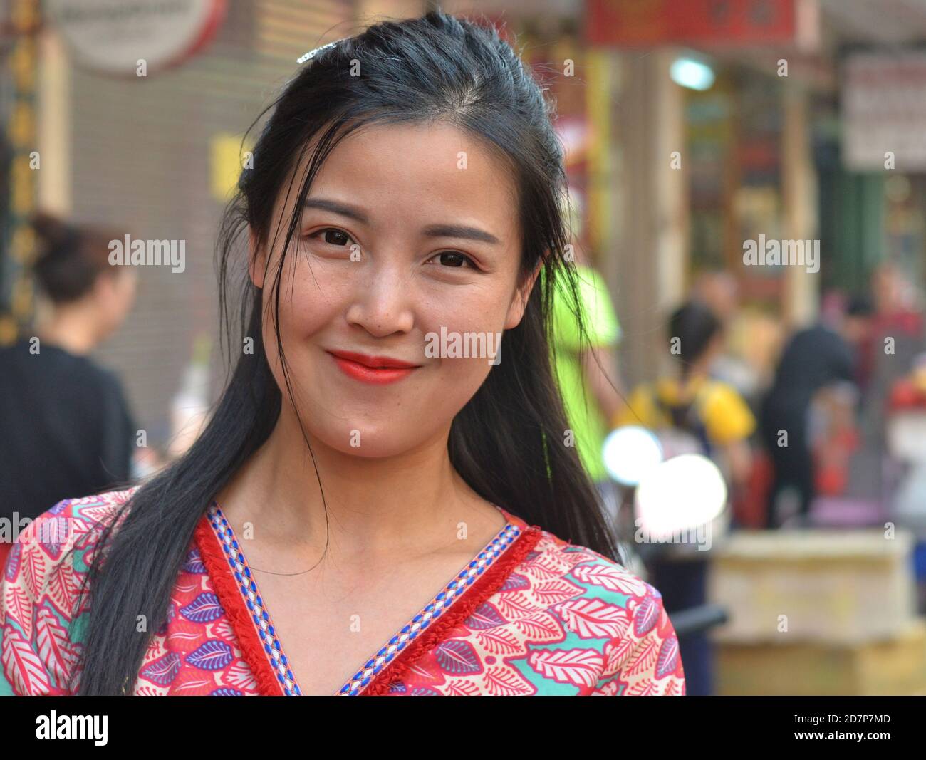 Closed-mouth smiling young South East Asian Thai woman with red lips and long hair poses for the camera on a busy street in downtown Bangkok. Stock Photo