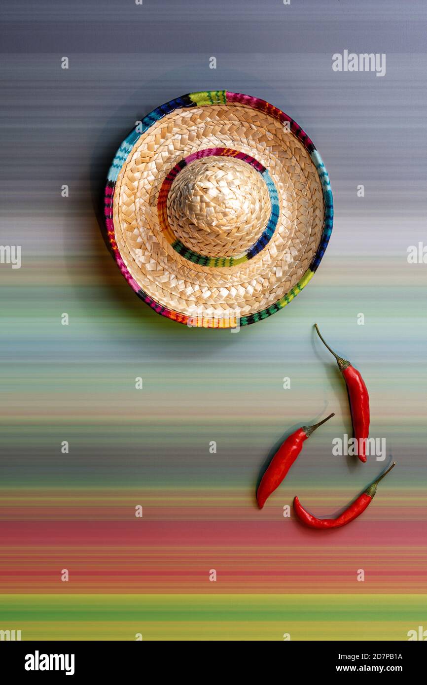 Colorful mexican straw hat next to three red hot chili peppers on a a colorful stripe pattern background. Mexico inspiration with copy space. Stock Photo