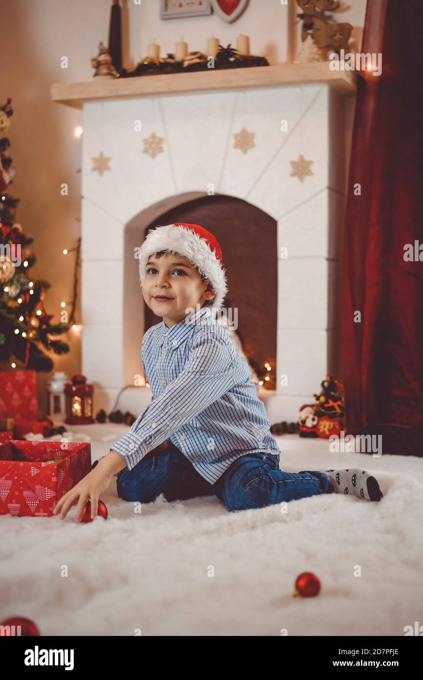 Kid with santa hat colecting Christmas tree balls and having fun in front of fireplace Stock Photo