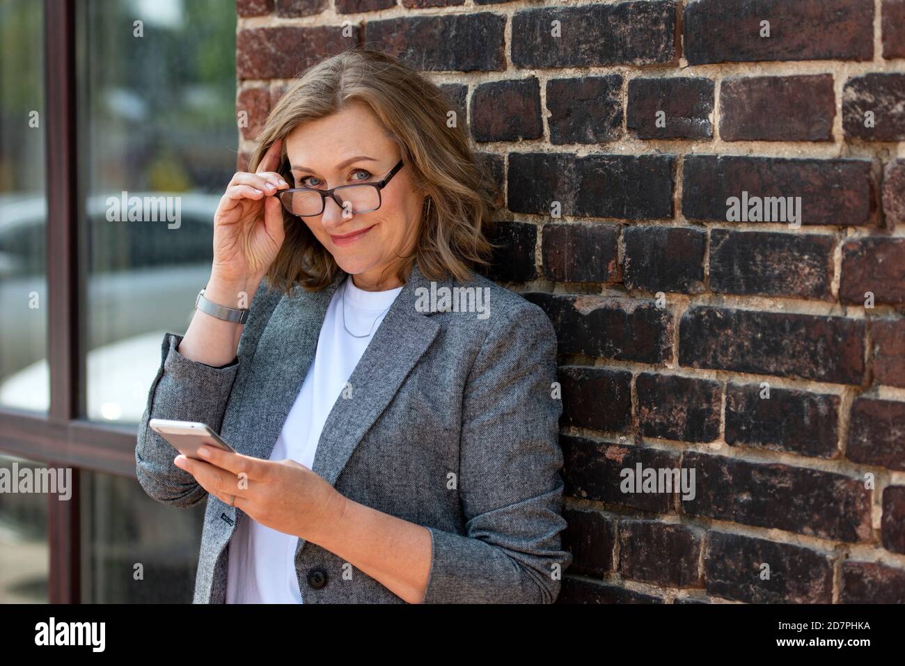 Portrait of a happy beautiful blonde woman, 55 years old, smiling, with a smartphone in hand. Outdoors, in the city. Stock Photo