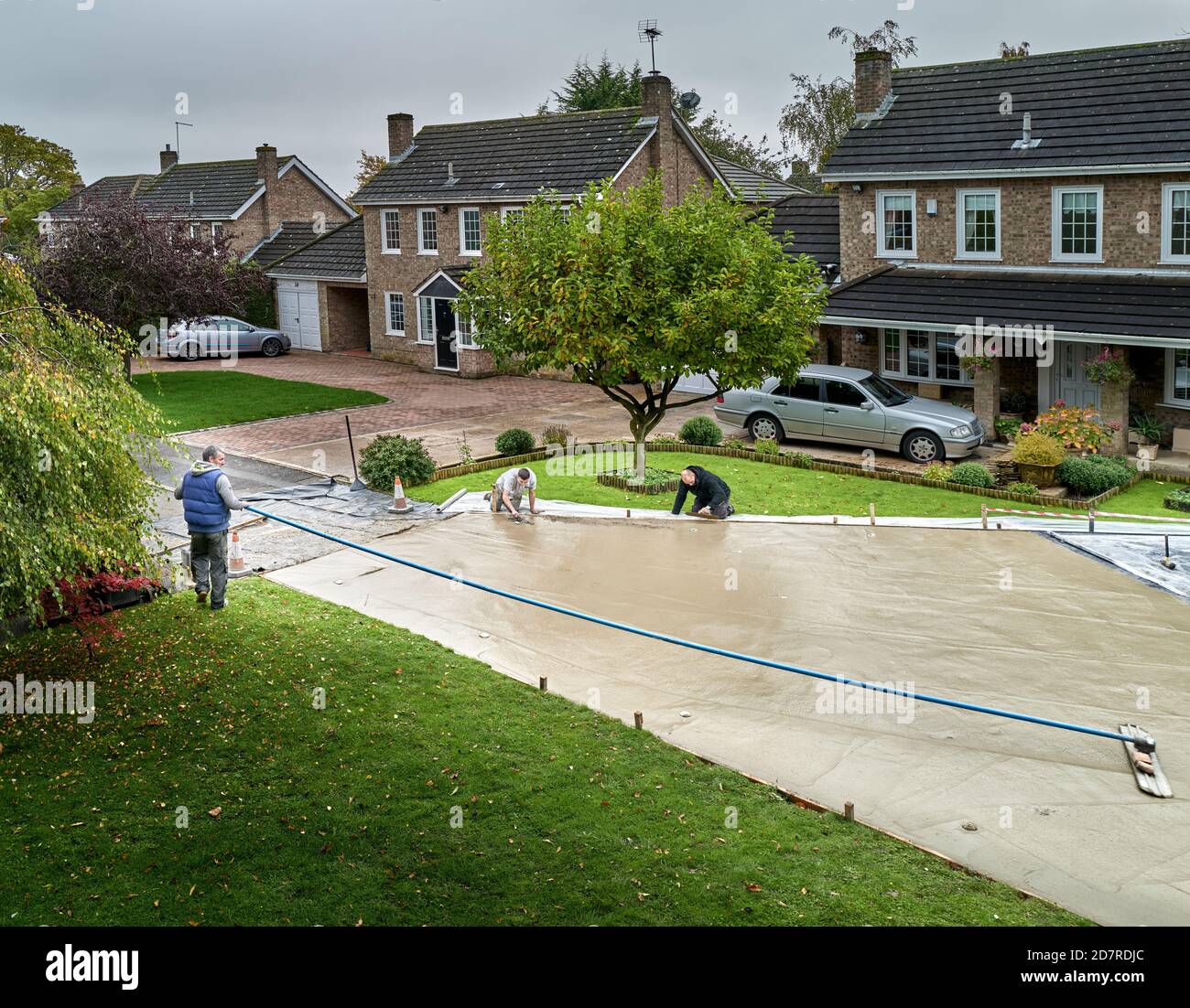 Workman with a long handled trowel screeds freshly laid concrete on a new driveway, whilst his two colleagues use short handled trowels. Stock Photo