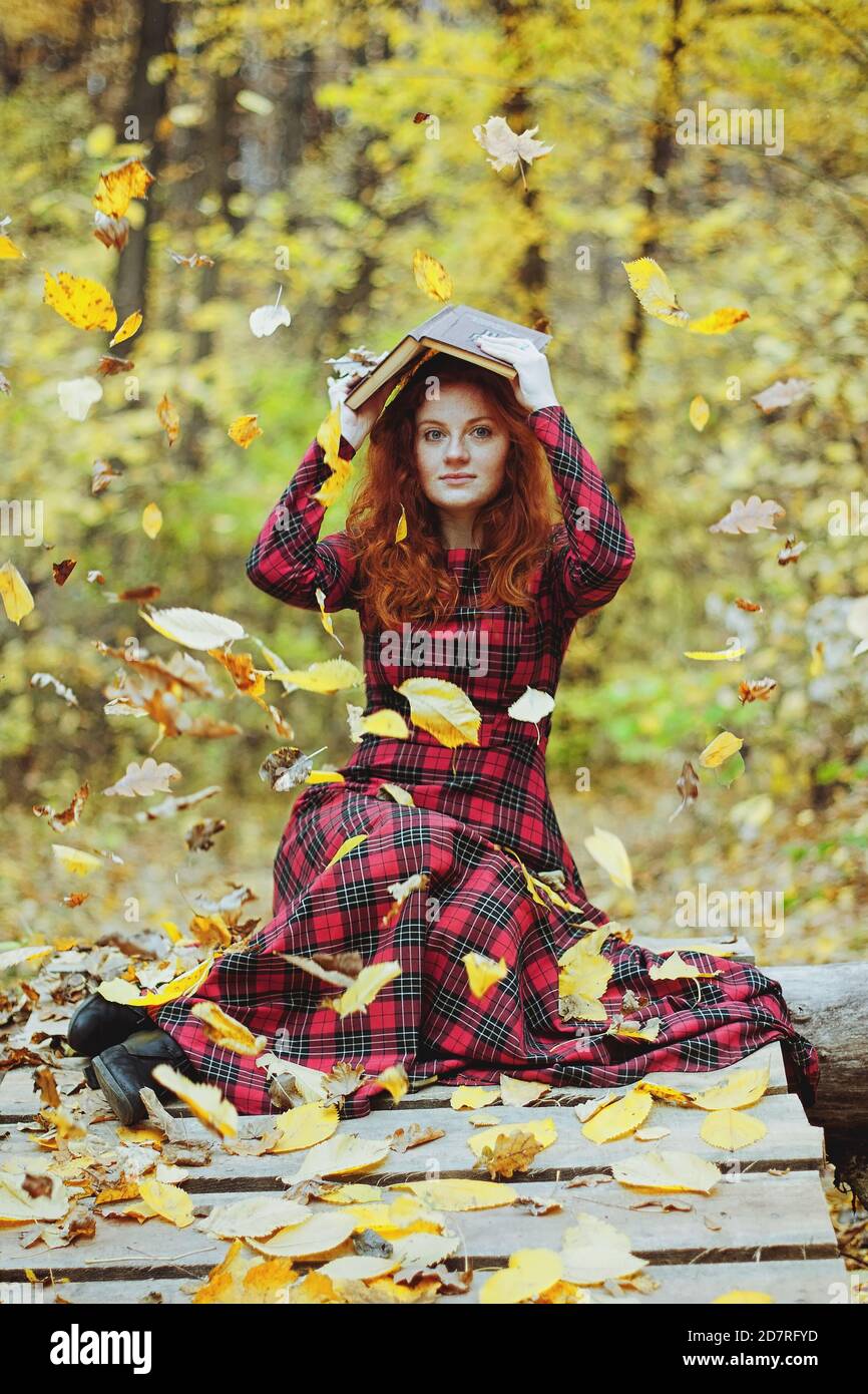 Happy girl with red hair and freckles in red dress reading a book in autumn park Stock Photo