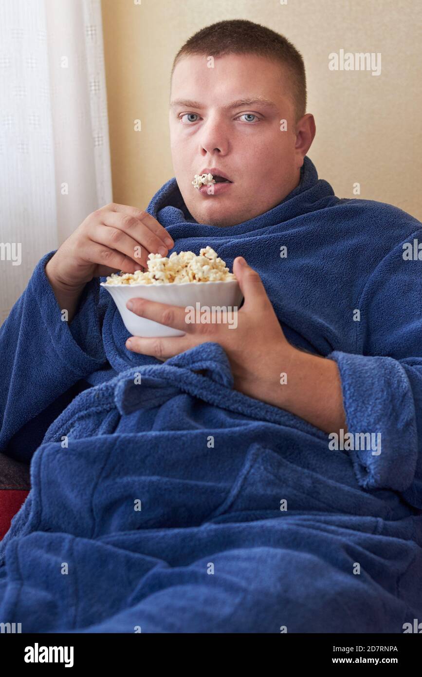 Fat man in a robe eating popcorn while sitting on the couch, obesity and improper lifestyle